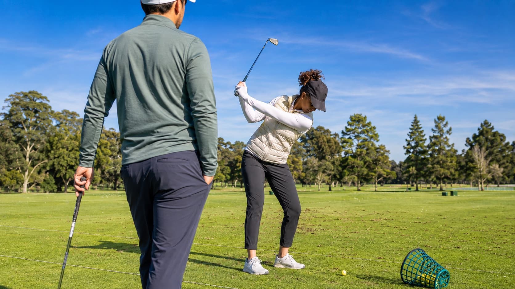 A golf coach watching a student practice her swing on a sunny driving range