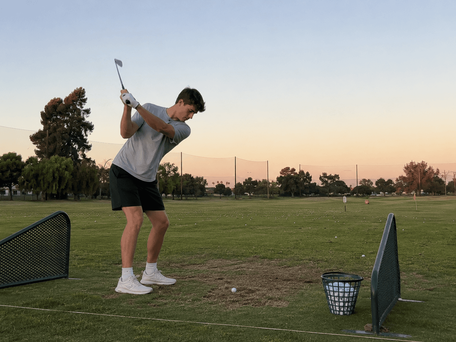 Young golfer practices alone at a local driving range in the early evening, working on his swing on a worn grass mat with a basket of balls nearby.