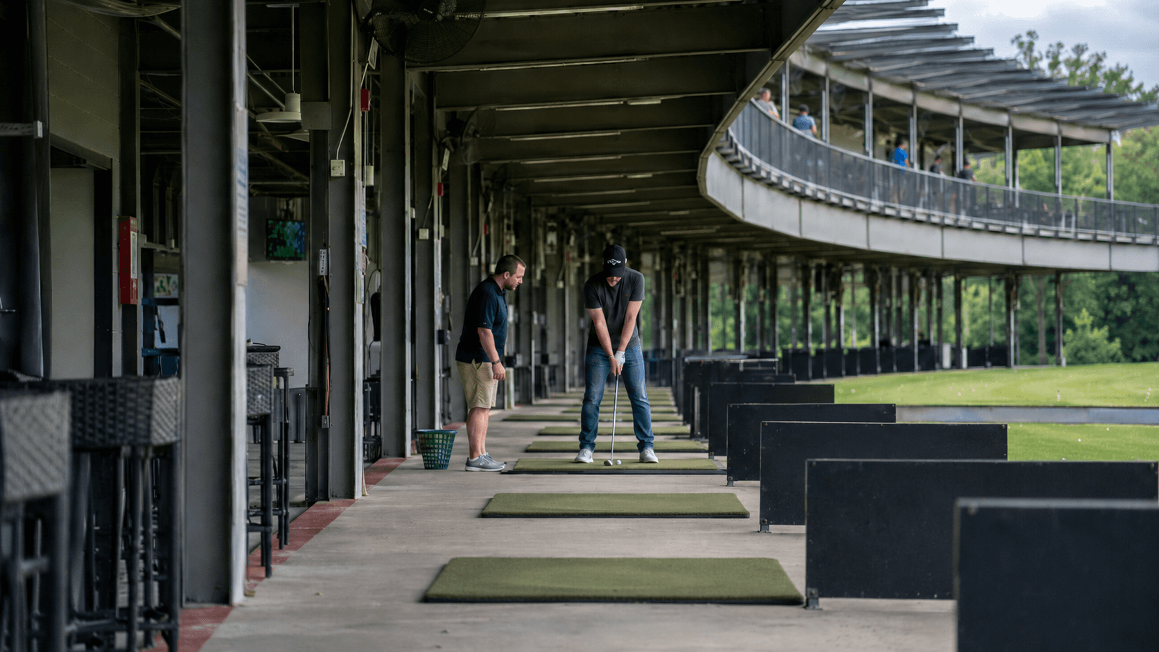 Golf coach watches a golfer set up to hit an iron from a mat at a two-level driving range.
