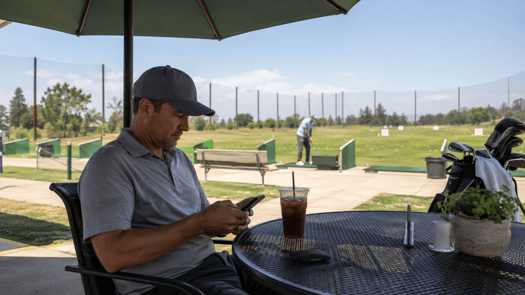 Golfer sits under a shade umbrella at an outdoor public golf course patio, looking at a phone with the driving range in the background.