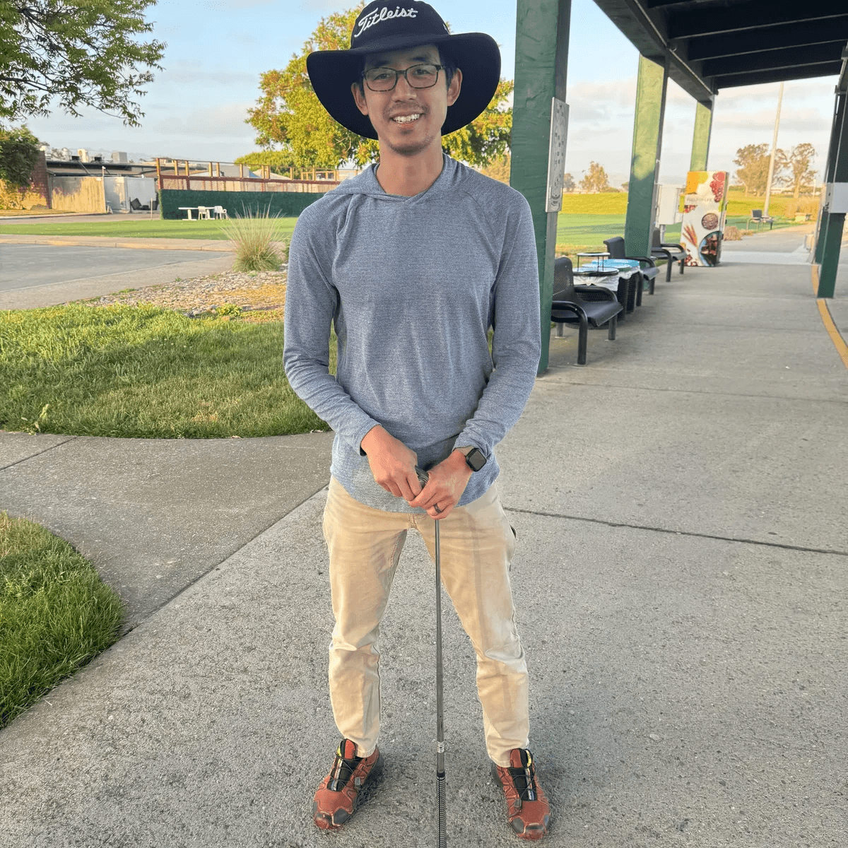 Larry Zhou holding a golf club outdoors, wearing a broad-brimmed hat and glasses.