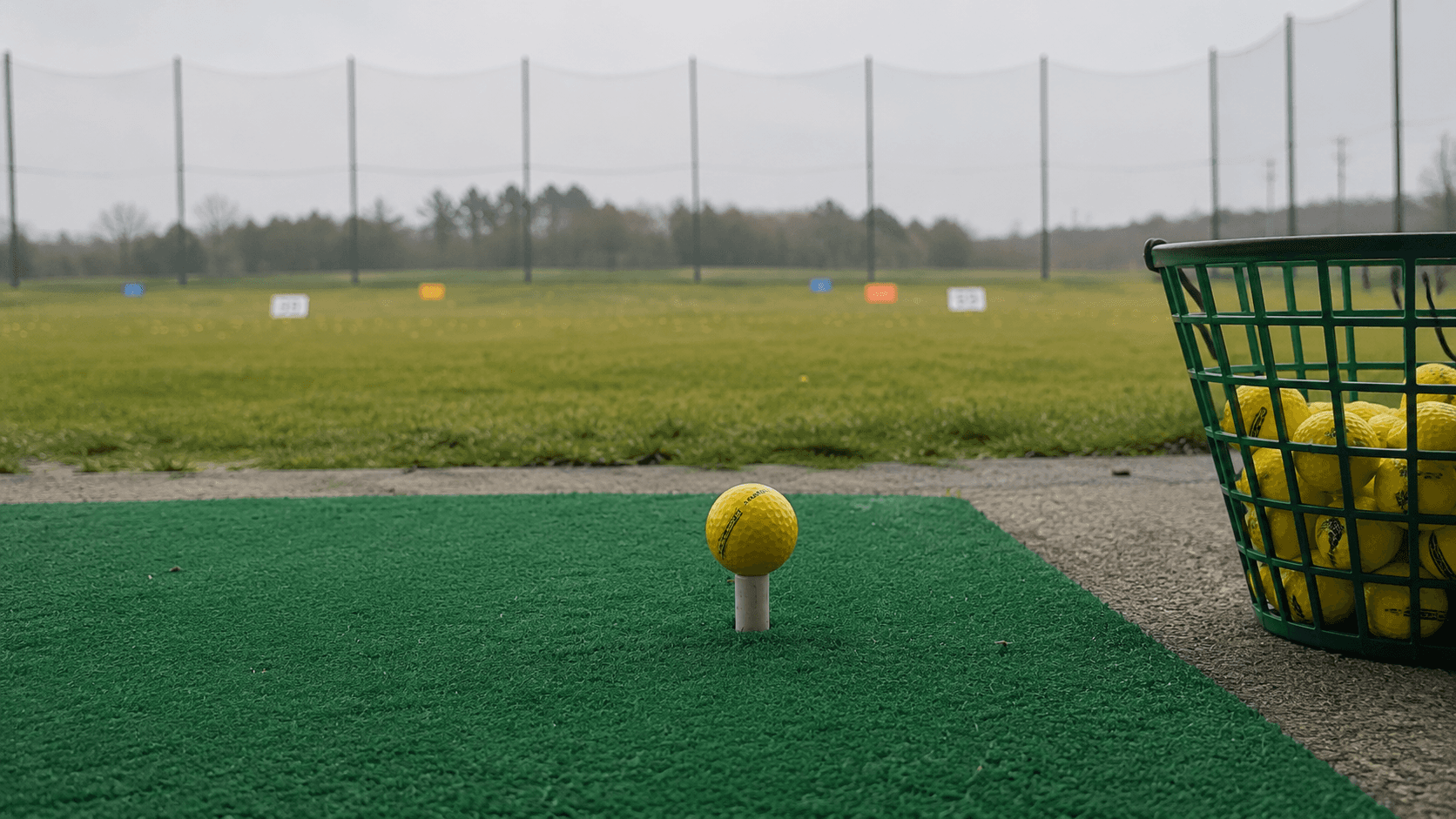 Yellow golf ball teed up on a white rubber tee beside a basket of range balls at an outdoor driving range.