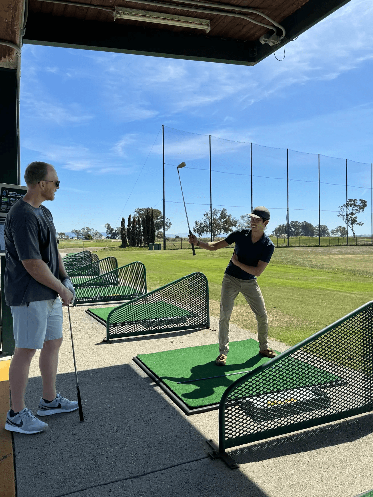 Larry Zhou demonstrates a golf swing while a student watches at a driving range.