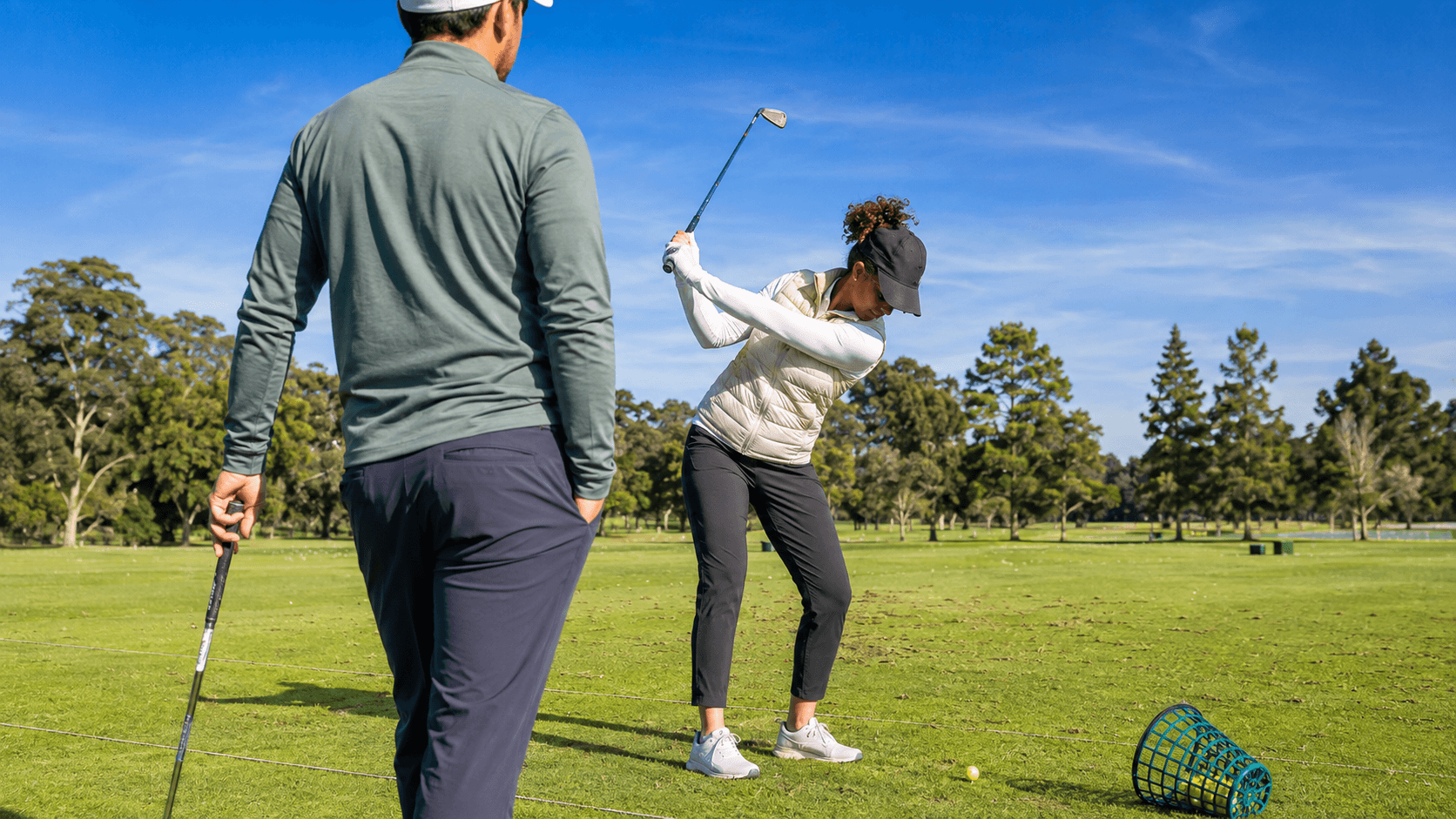 Golf coach watches a woman practice her backswing on an outdoor driving range with golf balls and a practice basket nearby.
