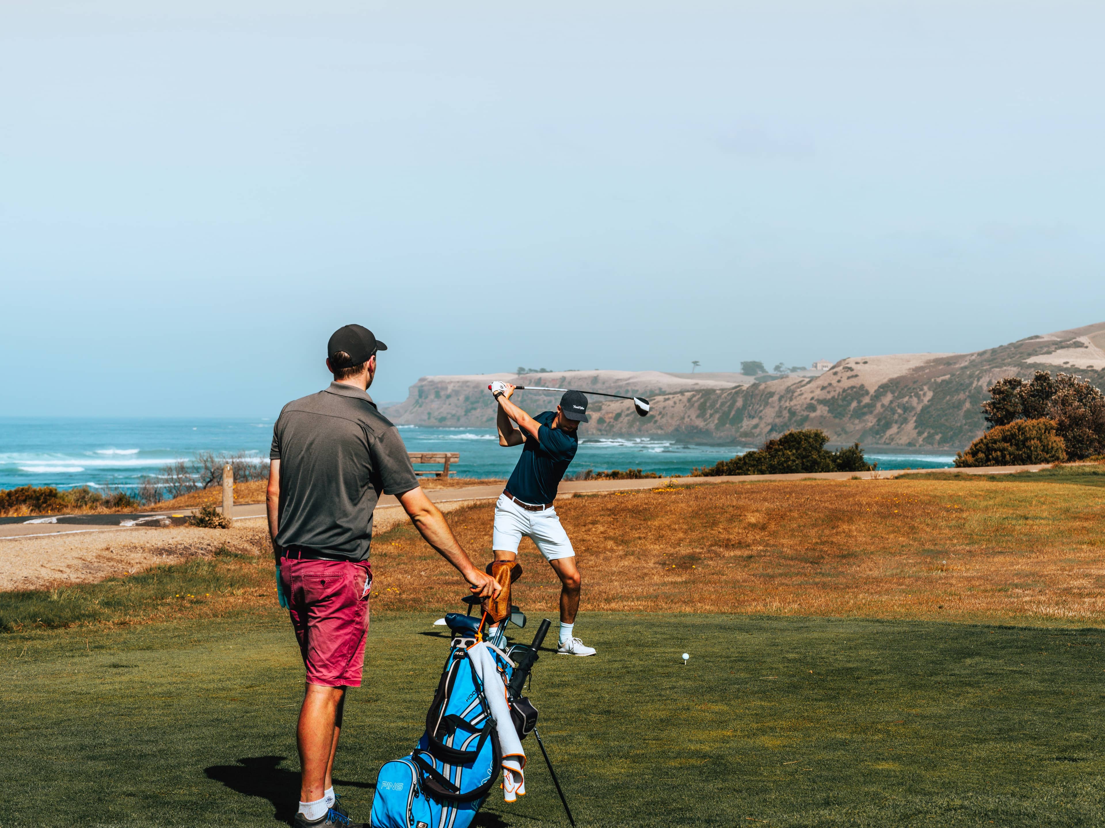 Man swinging a golf club on a course near the ocean while another man stands nearby watching.