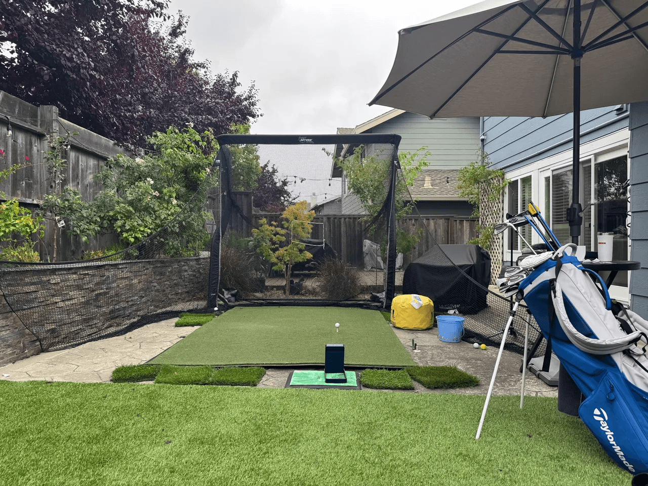 A golf practice net, hitting mat, launch monitor, and bag of clubs in an outdoor setup.