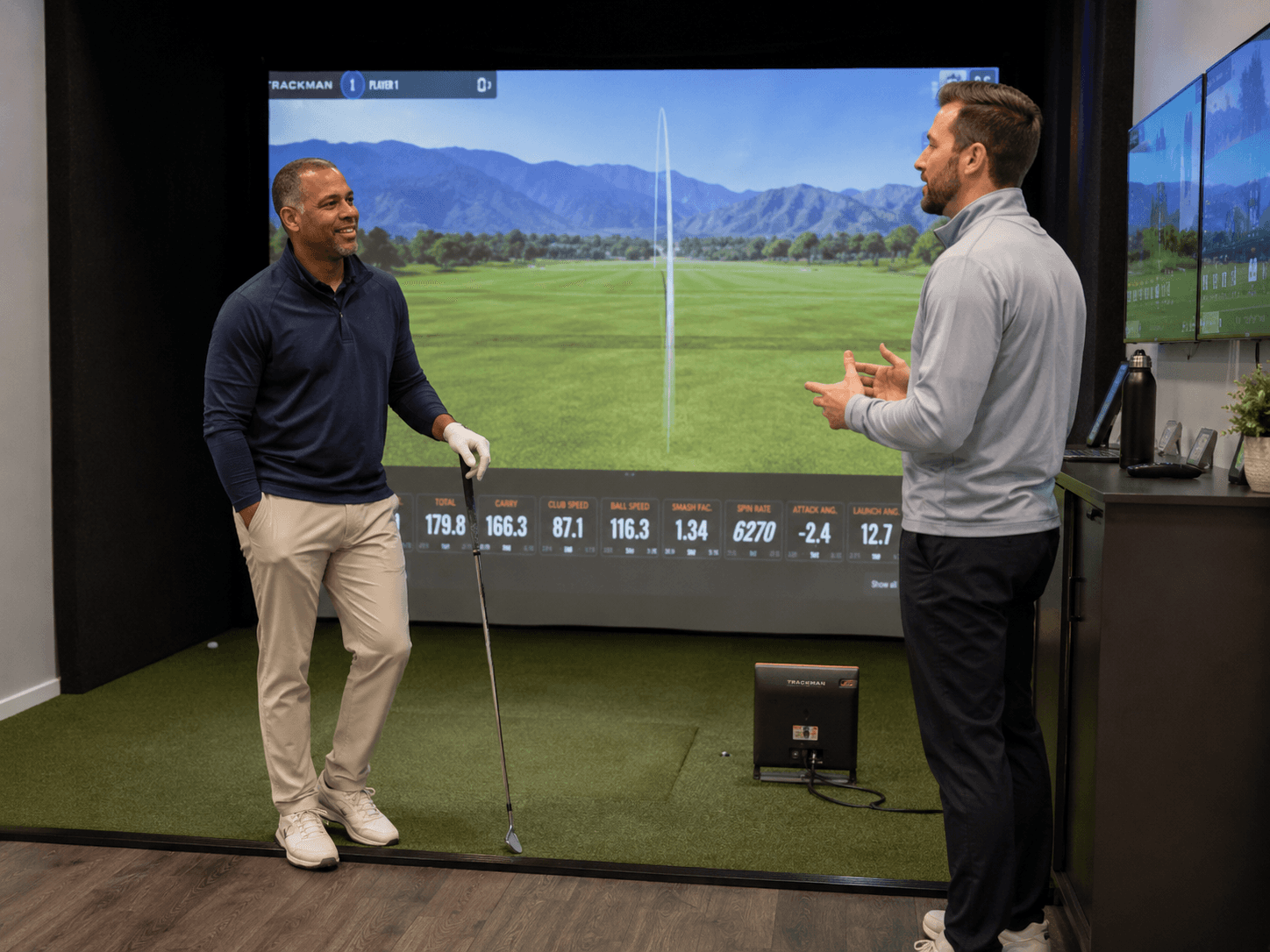 Golf coach speaking with an adult student holding a club on a hitting mat inside an indoor golf simulator studio before the start of a lesson.